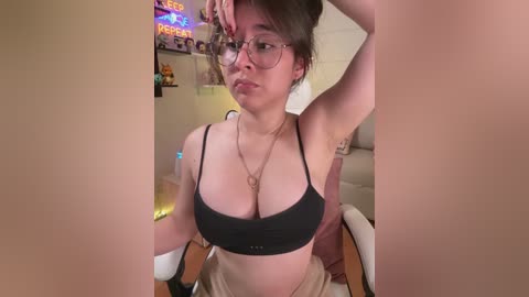 A video of a young woman with glasses, medium build, and medium-sized breasts wearing a black bra and beige pants, sitting in a dimly lit room with neon signs and gaming setup in the background.