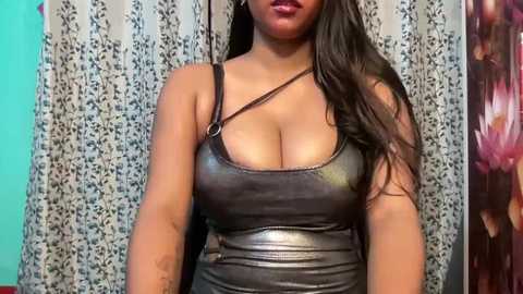 Video of a curvy, dark-skinned woman with long, wavy hair, wearing a revealing, black leather halter top, standing against a floral-patterned curtain.