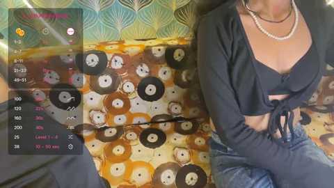 A video of a woman with a medium skin tone wearing a black crop top and pearls, sitting on a patterned couch. A smartphone screen shows a vinyl record player interface.