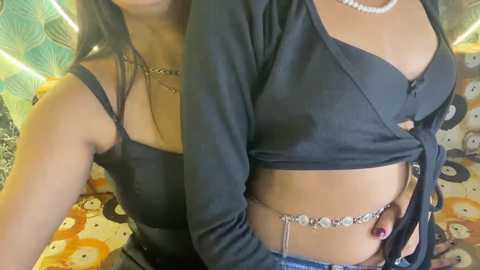 Video of two women, one with light skin and long dark hair, wearing a black, low-cut top, and the other with medium skin and medium-length dark hair, also in a black top. They are standing in a colorful, patterned room.