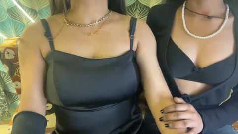 Video of two women, both wearing black tops and pearl necklaces, embracing indoors with patterned green walls in the background.