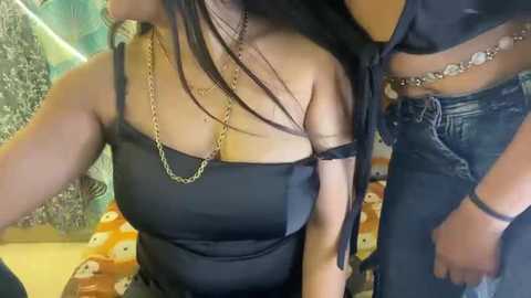 Video of two women in casual attire; one in a black off-shoulder top, chain necklace, and long dark hair; the other in blue jeans with a silver belt. Background features patterned wall and a table with cartoon-themed placemat.