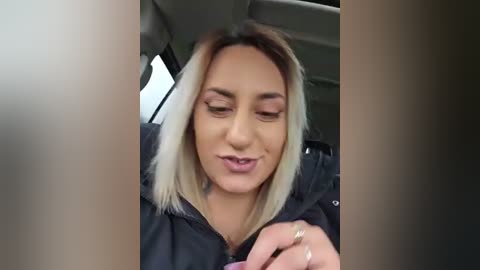 Video of a woman with blonde hair in a car, wearing a black jacket, smiling, and holding a ring, with blurred car interior in background.
