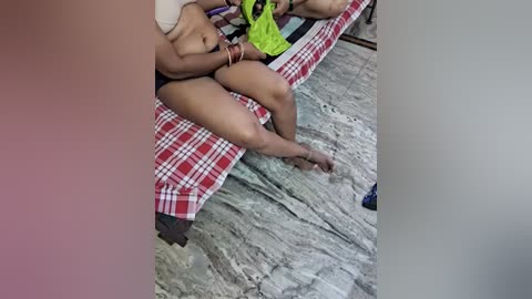 Video of a nude woman with medium brown skin, small breasts, and black hair, lying on a bed with a red and white checkered blanket, legs spread, holding a green shirt. Background shows tiled floor and partially visible bathroom.