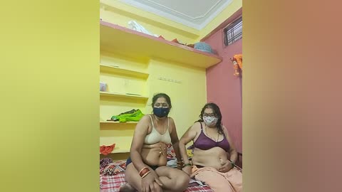 Video of two women in casual clothing, seated on a bed in a small, brightly colored room with yellow walls and a pink accent wall. The women wear face masks and have diverse body types.