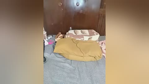 Video of a messy bedroom with a brown blanket, pink pillow, stuffed animal, and scattered clothes on a gray bed, against a wooden wardrobe backdrop.