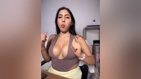Video of a Latina woman with long black hair, wearing a low-cut brown top revealing cleavage, sitting at a desk, making a playful gesture with her hands.