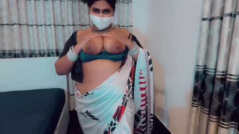 Video of a South Asian woman with medium skin tone, dark hair, and medium breasts. She is wearing a white saree, a green bra, and a face mask, lifting the saree to reveal her breasts. Background includes a bed and curtains.