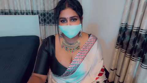 Video of a South Asian woman in a black blouse and white saree with red border, wearing a blue surgical mask, gold jewelry, and sitting indoors with beige curtains.