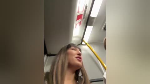 A video of a smiling Asian woman with straight, shoulder-length brown hair, standing in a subway car, holding a pole with yellow handles. The background shows white subway walls and fluorescent lights.