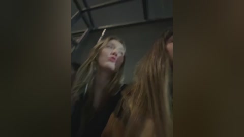 Video of two women in a dimly lit room, one with light brown hair and a black top, the other with long, wavy blonde hair, both appearing tense and talking.