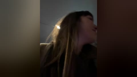 A dimly lit video of a woman with long, straight brown hair, wearing a dark coat, leaning against a wooden surface, her head tilted slightly to the left.