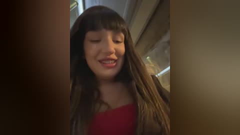 Video of a young, light-skinned Asian girl with long, straight brown hair and bangs, wearing a red top, smiling broadly, indoors with blurred background.