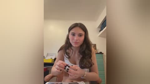 Video of a young woman with long brown hair, wearing a white tank top, sitting at a table. She holds a small, black object in her hands. The background features a cluttered room with white walls and a window.