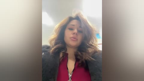Video of a woman with wavy, shoulder-length brown hair, wearing a red blouse and a dark jacket, indoors with blurred background.