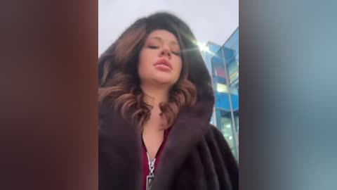 Media: Video of a young woman with wavy, shoulder-length brown hair, wearing a dark coat and red top. She has a closed-mouth smile, standing in front of a glass building with a blurred background.