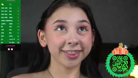 A video of a smiling young woman with long black hair, light skin, and dark eyes, wearing a nose ring. The background shows a green digital overlay with numbers and icons.