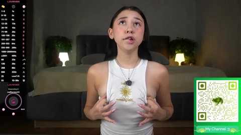 Video of a young woman with long black hair, wearing a white tank top, standing in a dimly lit bedroom with a bed, potted plants, and a TV showing a green QR code.