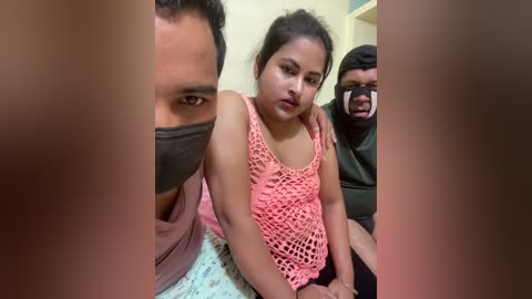 Video of a young South Asian woman with a light complexion, wearing a pink crocheted top, sitting on a bed, surrounded by three men in masks, one holding her shoulder.