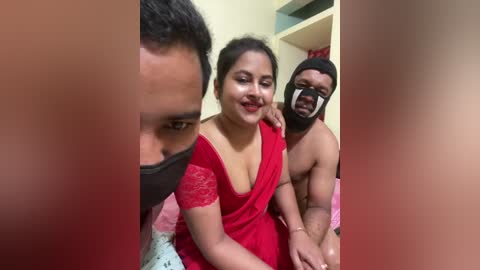Video of a woman in a red sari, a man wearing a mask, and another man shirtless, all indoors with blurred faces.