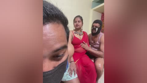 Video of a woman in a red dress with a man in a black mask, sitting on a sofa in a domestic setting, blurred faces, neutral expression.