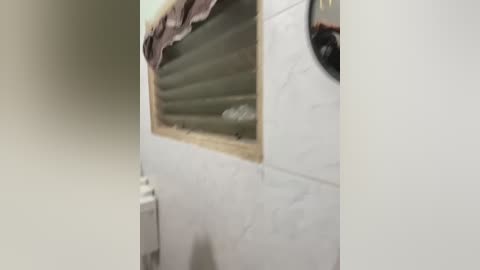 A video of a white-tiled bathroom wall with a partially visible, narrow window. The window has a light-colored blind and a pink cloth draped over it. The tiles are clean and glossy, reflecting light.