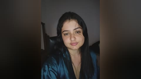 A video of a young woman with long black hair, light skin, and a neutral expression, wearing a blue satin robe, seated in a dimly lit room with dark furniture.