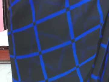 Video of a person wearing a black shirt with a vibrant blue grid pattern, seen from the waist up. Background shows a pink wall and a wooden bench.