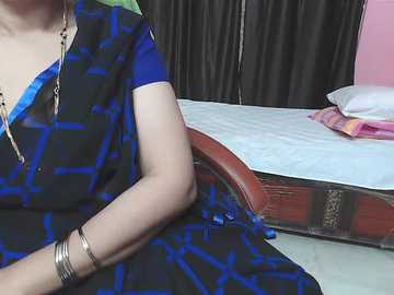 Video of a woman in a blue-patterned sari, seated on a bed with a pink pillow, against black curtains.