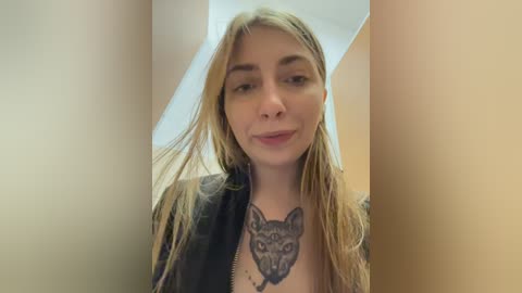 Video of a young, fair-skinned woman with long, straight blonde hair, wearing a black jacket and revealing a detailed, black wolf tattoo on her chest. She has a subtle smile, and the background is blurred, focusing on her face.