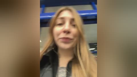 A video of a young Caucasian woman with long blonde hair, wearing a black jacket over a grey sweater, smiling slightly. Background shows a blue ceiling and blurred, indistinct objects.