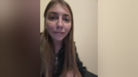 A blurry video of a young woman with long, light brown hair, wearing a black jacket, standing against a beige wall in a dimly lit room.