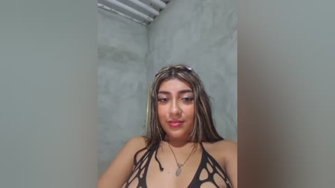 A video of a young Latina woman with long, dark hair, wearing a black, strappy, cut-out swimsuit, standing in a dimly lit, concrete-walled room with a metal ceiling.