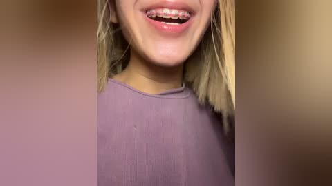 Video of a close-up of a smiling young woman with light skin, straight blonde hair, and braces, wearing a purple shirt, against a blurred background.