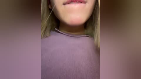 Video of a young woman with fair skin, straight blonde hair, and light pink lips, wearing a tight, ribbed purple top. The background is a blurred, neutral-toned wall.