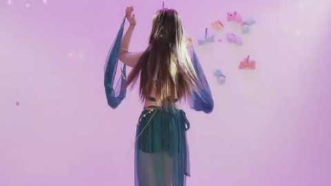 Video of a woman with long blonde hair, wearing a teal flowing dress and sheer blue sleeves, dancing against a pink background adorned with floating pink and purple balloons.