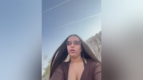 Video of a young woman with long brown hair, wearing brown sunglasses, a sheer brown top, and a large chest, standing in front of a blurred, beachy background.
