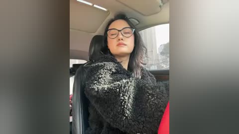 Video of an East Asian woman with glasses, long black hair, wearing a black fur coat, leaning back in a car seat with a neutral expression.