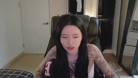 Video of a young Asian woman with long black hair, wearing a beige sweater, sitting on a black leather chair in a modern, minimalist room with beige walls, white door, and a closet.
