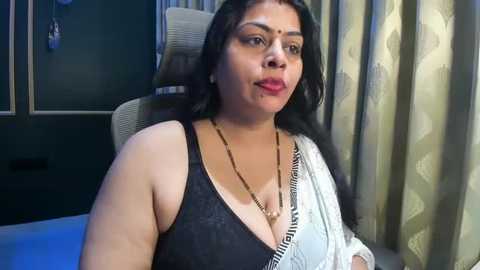 Video of a South Asian woman with medium skin tone and long black hair, wearing a black lace bra and a white saree, sitting on a grey chair with green curtains in the background.