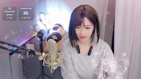 A video of a young East Asian woman with straight black hair, wearing a light grey top, recording herself on a professional microphone.
