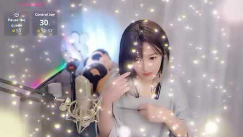 A video of an Asian woman with straight black hair, wearing a gray top, sitting in front of a microphone, singing. The background is blurred with sparkling particles.
