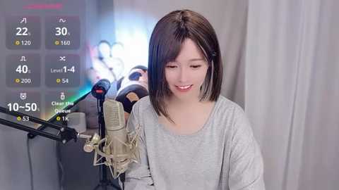 Video of a young Asian woman with straight black hair, wearing a grey long-sleeve shirt, singing into a microphone, with a studio background displaying temperature and humidity levels.