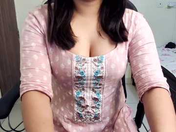 Video of a woman with light brown skin, medium build, wearing a pink top with a decorative blue floral panel, sitting at a desk in a room with a wooden door and white walls.