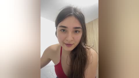 A video of a young woman with fair skin, dark hair, and brown eyes, wearing a red spaghetti-strap top, standing in a brightly lit room with white walls and wooden paneling.