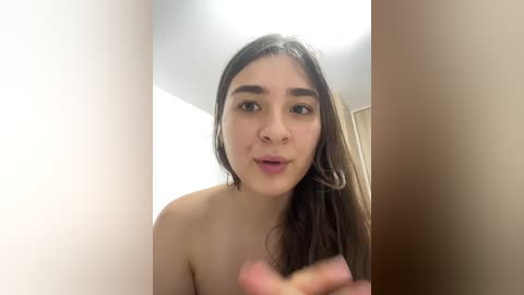 Video of a young, light-skinned woman with long, dark brown hair, slightly parted lips, and bare shoulders. She is indoors, facing the camera with a neutral expression.