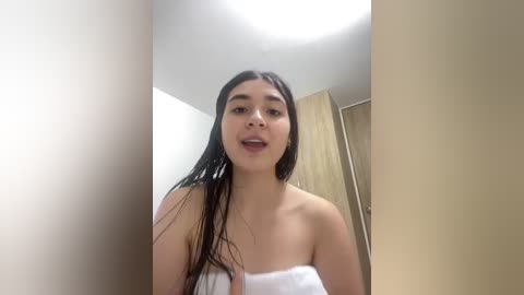 A video of a young Asian woman with long black hair, wet from a shower, wearing a white towel, standing in a beige-tiled bathroom with a wooden door in the background.