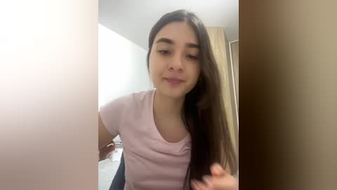 Video of a young woman with long dark hair, wearing a light pink t-shirt, sitting in a brightly lit room with beige walls.