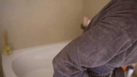 Video of a person in a gray bathrobe, standing in a beige-tiled bathroom. A white bathtub is partially visible in the background, with a bottle of shampoo on the edge.