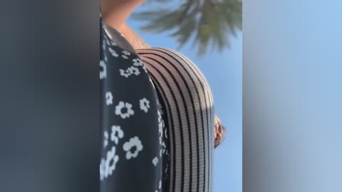 Video of a person's lower back and buttocks, wearing a striped, see-through, black and white dress, against a clear blue sky and a palm tree in the background.
